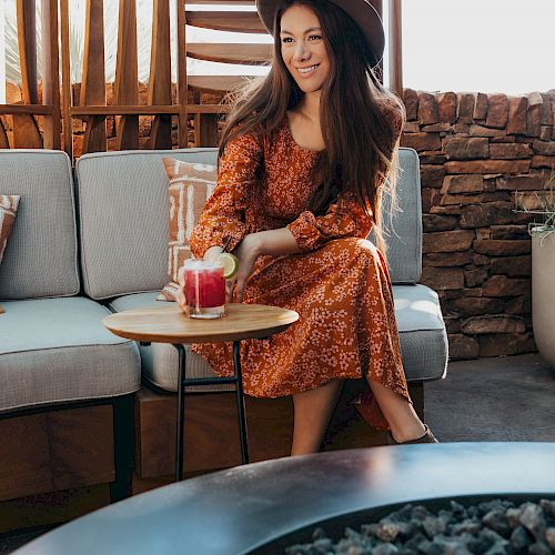 A woman in a hat and floral dress sits on a couch, reaching for a drink on a small table in a cozy outdoor setting.