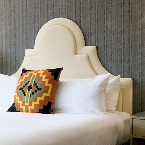 A modern bedroom with a white bed, a patterned pillow, two side tables, and two lamps, featuring a cushioned headboard and textured wall.