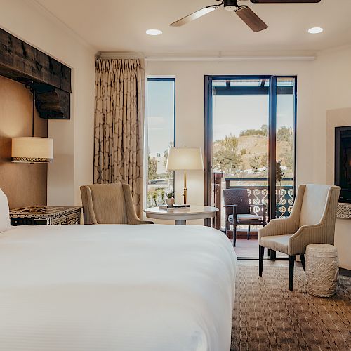 This image shows a cozy hotel room with a bed, chairs, a dresser, and a balcony overlooking greenery, featuring stylish décor and a warm ambiance.