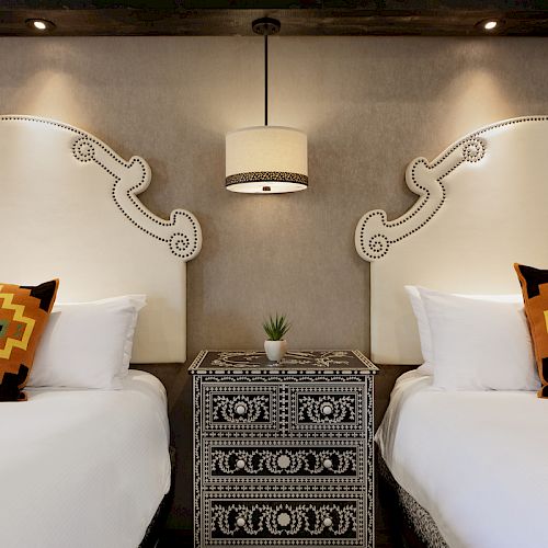 The image shows two beds with white sheets, colorful patterned pillows, a patterned bedside table with a plant, and a pendant light above.