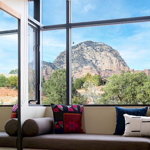 A cozy seating area with colorful pillows overlooks a stunning mountain view, framed by large windows, with a patterned rug on the wooden floor.