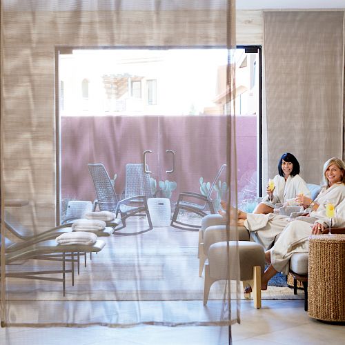 Three people in robes are relaxing and chatting in a spa lounge area, with lounge chairs and a sunny outdoor space visible through a window.