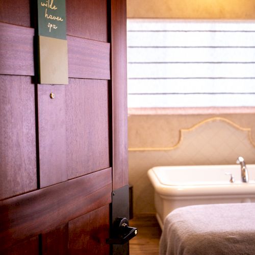 An open door reveals a room with a bathtub, a window with blinds, and a table covered with a towel, suggesting a serene, spa-like setting.