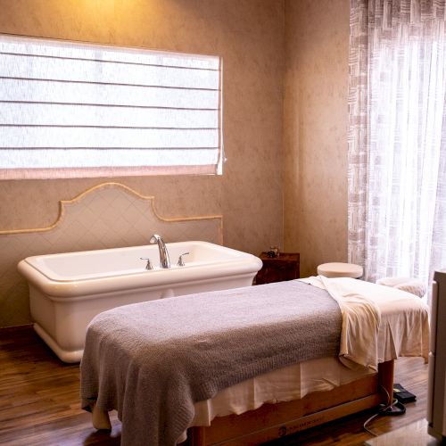 The image shows a cozy spa room with a massage table covered in towels and a bathtub under a window with blinds, next to a curtained glass door.