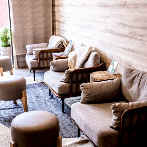 The image shows a cozy seating area with beige armchairs and stools, a wooden wall, and soft carpet, creating a warm and inviting atmosphere.