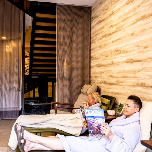 Two people are relaxing on lounge chairs in a cozy room, wearing white robes. One is reading a magazine, and the room has a warm and inviting ambiance.