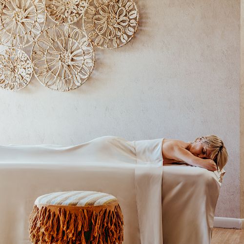 A person is lying on a massage table covered with a white sheet in a room decorated with woven wall hangings and a cushioned stool.