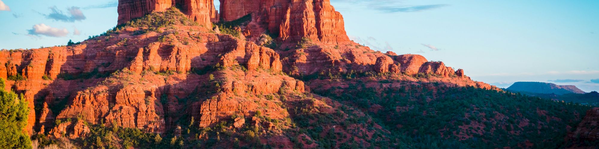 The image shows a stunning red rock formation bathed in sunlight, likely in a desert or canyon setting, with shrubs and trees in the foreground.