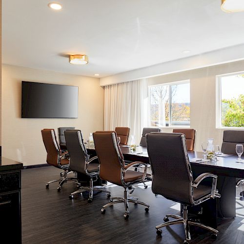 A modern conference room with a long table, eight chairs, a wall-mounted TV, large windows, and a side counter with items on it.