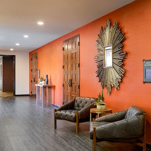 A modern hallway with an orange accent wall, decorative mirror, two brown chairs with a small table, and wooden doors on both sides ends the sentence.