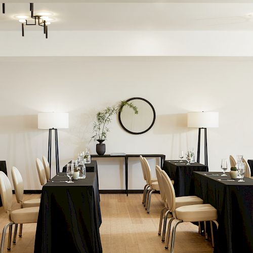 A small conference room with tables, chairs, black tablecloths, wine glasses, and a decorative wall mirror. The room has modern lighting and minimal décor.