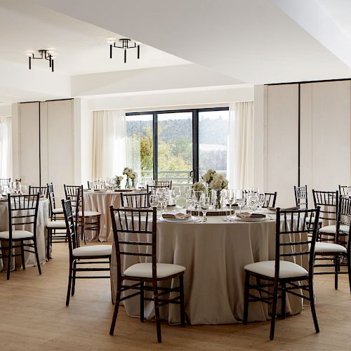 The image shows a banquet hall with round tables set for an event, each adorned with white cloths, floral centerpieces, and surrounded by black chairs.