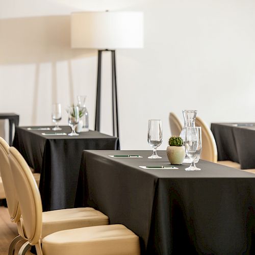 A modern indoor setting with tables draped in black tablecloths, beige chairs, wine glasses, a floor lamp, and small plants, creating an elegant atmosphere.