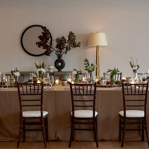 A long dining table set for a formal meal with brown chairs, elegant table settings, floral decorations, and a round mirror and lamp in the background.