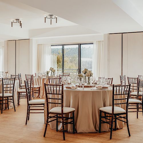 A beautifully decorated banquet hall with round tables, chairs, floral centerpieces, and large windows offering natural light, ready for an event.