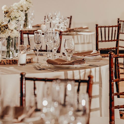 The image shows an elegantly set event table with white floral arrangements, multiple wine glasses, napkins, and wooden chairs around.