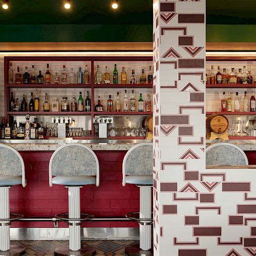 The image shows a stylish bar with patterned tiles, stocked shelves of bottles, and three tall bar stools, featuring a geometric design on the column.