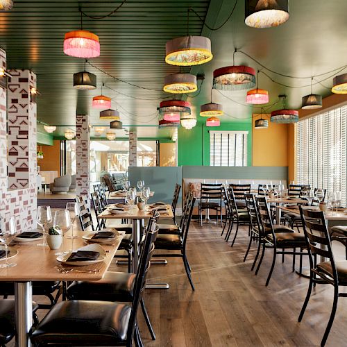 A colorful, modern restaurant with empty tables, chairs, and vibrant hanging lamps. Large windows let in natural light, creating a bright atmosphere.