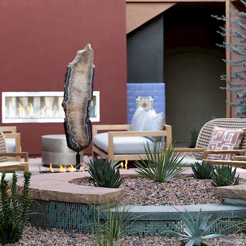 Outdoor seating area with modern chairs, a fire pit, and low vegetation; a structure in the background with a wall-mounted fireplace.