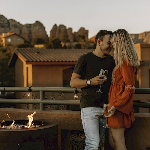 Two people stand close together on a terrace, holding drinks. There's a fire pit, and the background has houses and rocky hills at sunset.