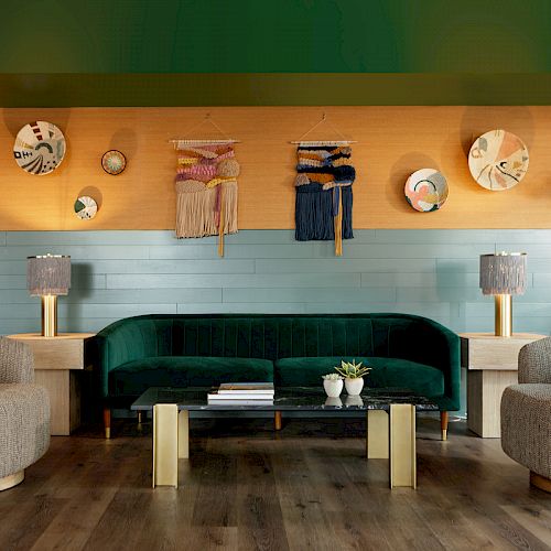 A stylish living room with a green sofa, two beige chairs, a black coffee table, and decorative wall art. Two lamps and potted plants are present.