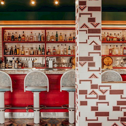 A colorful bar interior with a patterned column, three bar stools, a red bar counter, and shelves lined with various bottles in the background.