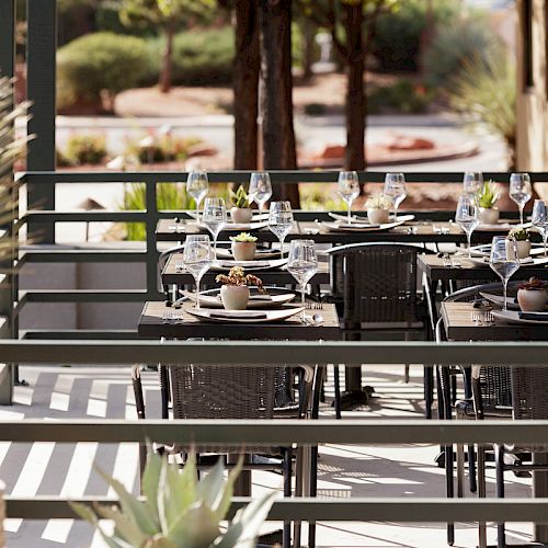 An outdoor dining area set with tables, chairs, plates, cutlery, and wine glasses, surrounded by plants and a sunny, tree-lined background.