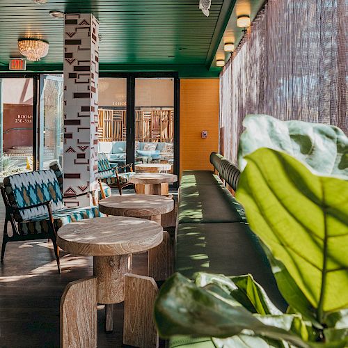 A cozy indoor seating area with green accents, wooden tables, patterned pillows, and a large leafy plant by the window, providing a relaxing atmosphere.