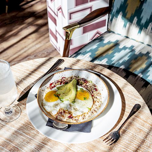 A table setting with a plate of food, featuring avocado and eggs, a glass of water, and cutlery on a round wooden table next to a cushioned chair.