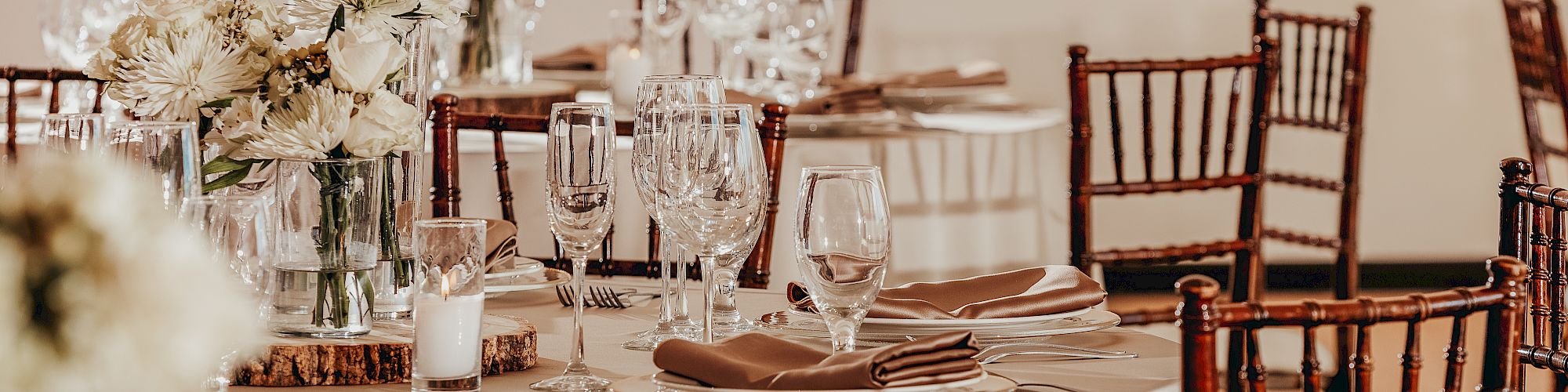 The image shows an elegant dining setup with round tables, wooden chairs, white floral centerpieces, and various glassware, ready for a formal event.