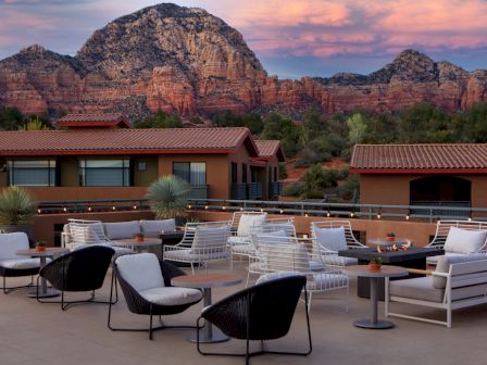 A rooftop seating area with modern furniture overlooks a stunning landscape of red rock mountains under a colorful sky at sunset.