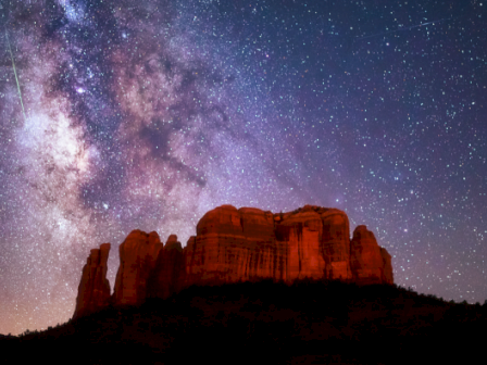 A stunning night sky with the Milky Way and shooting stars above dramatic rock formations, illuminated by soft, reddish light.