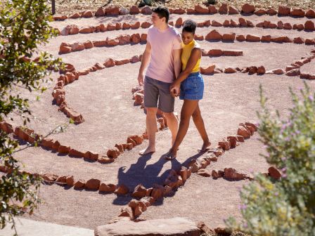 A couple is walking together on a dirt path surrounded by arranged rocks, holding hands, and enjoying an outdoor activity in a sunny setting.