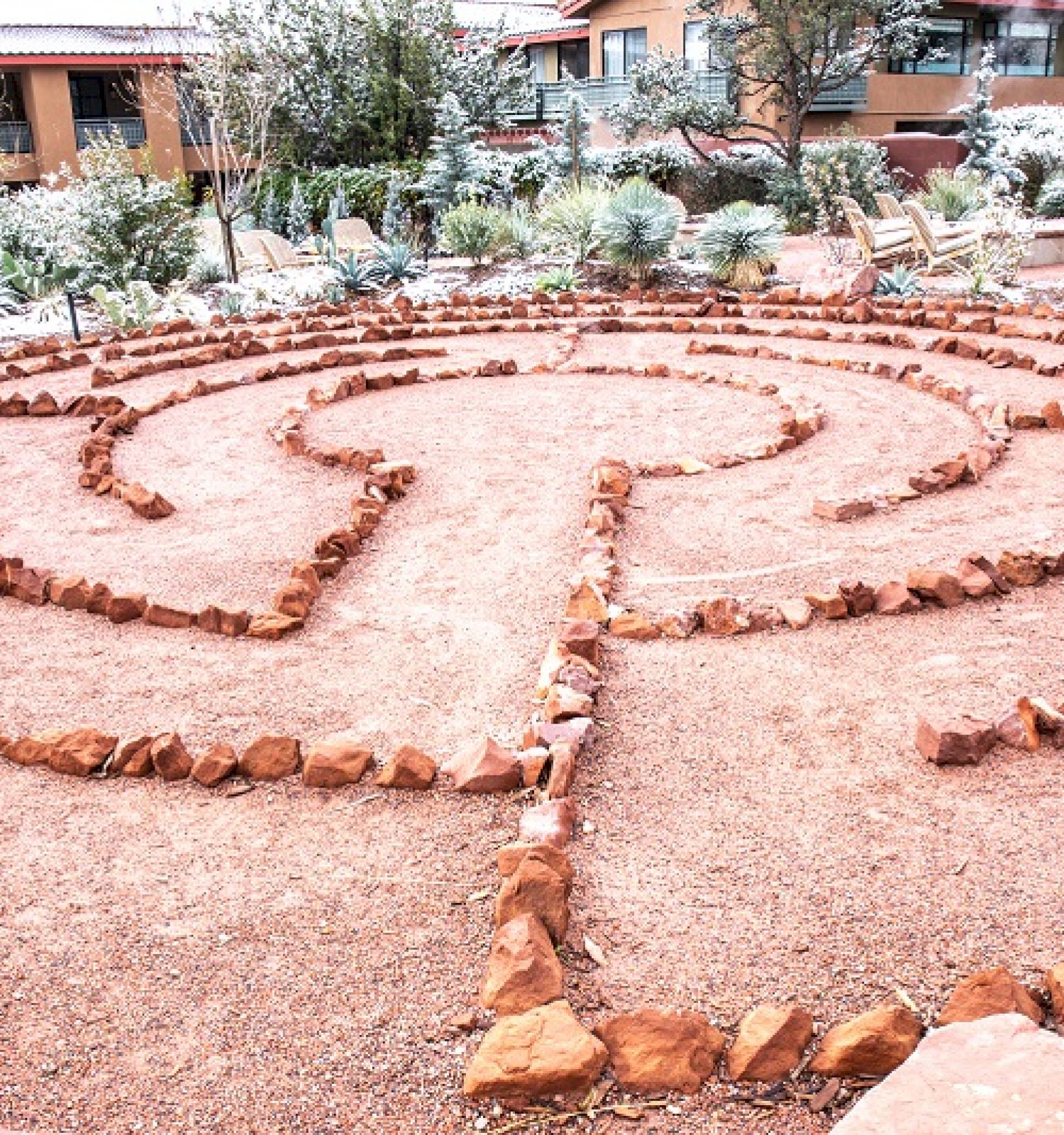 A garden with a labyrinth made of stones surrounded by light snow in a residential area with buildings and greenery in the background.