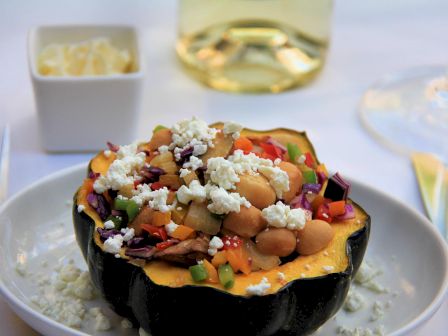 Stuffed acorn squash with beans, vegetables, and crumbled cheese, served on a white plate, with a side of butter, wine, and cutlery.