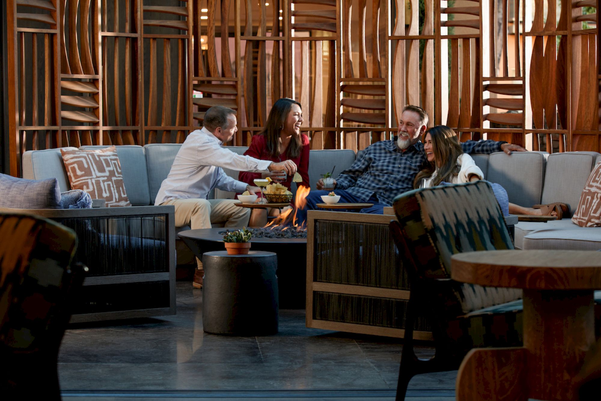 Four people are sitting on couches around a table, enjoying snacks and drinks, with a decorative wooden backdrop behind them.