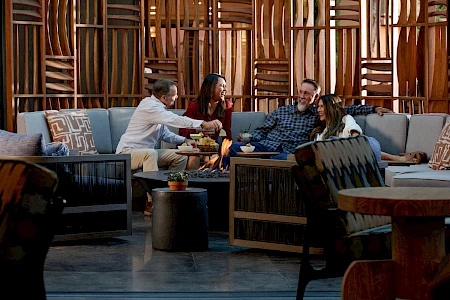 Four people are sitting on couches around a table, enjoying snacks and drinks, with a decorative wooden backdrop behind them.