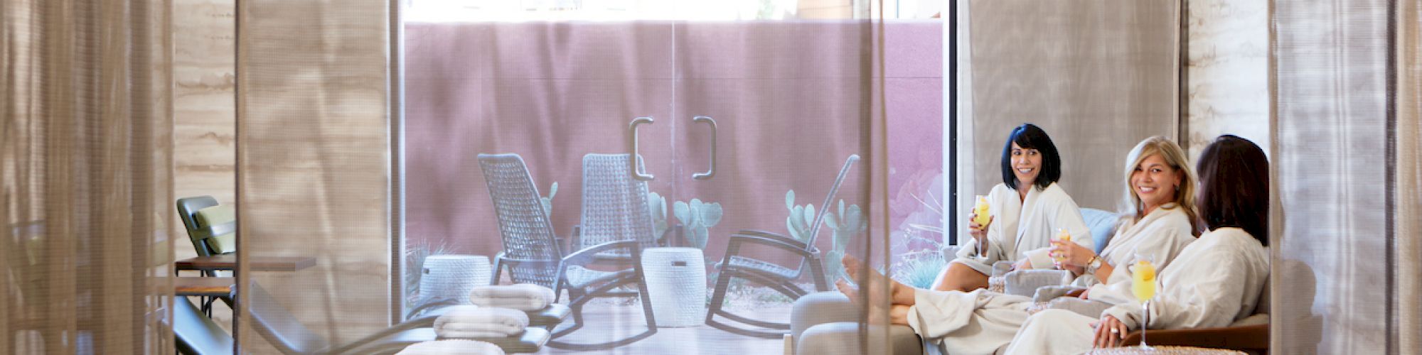 Three people in bathrobes relax indoors with drinks, while lounge chairs can be seen outside through a large window covered by a sheer curtain.