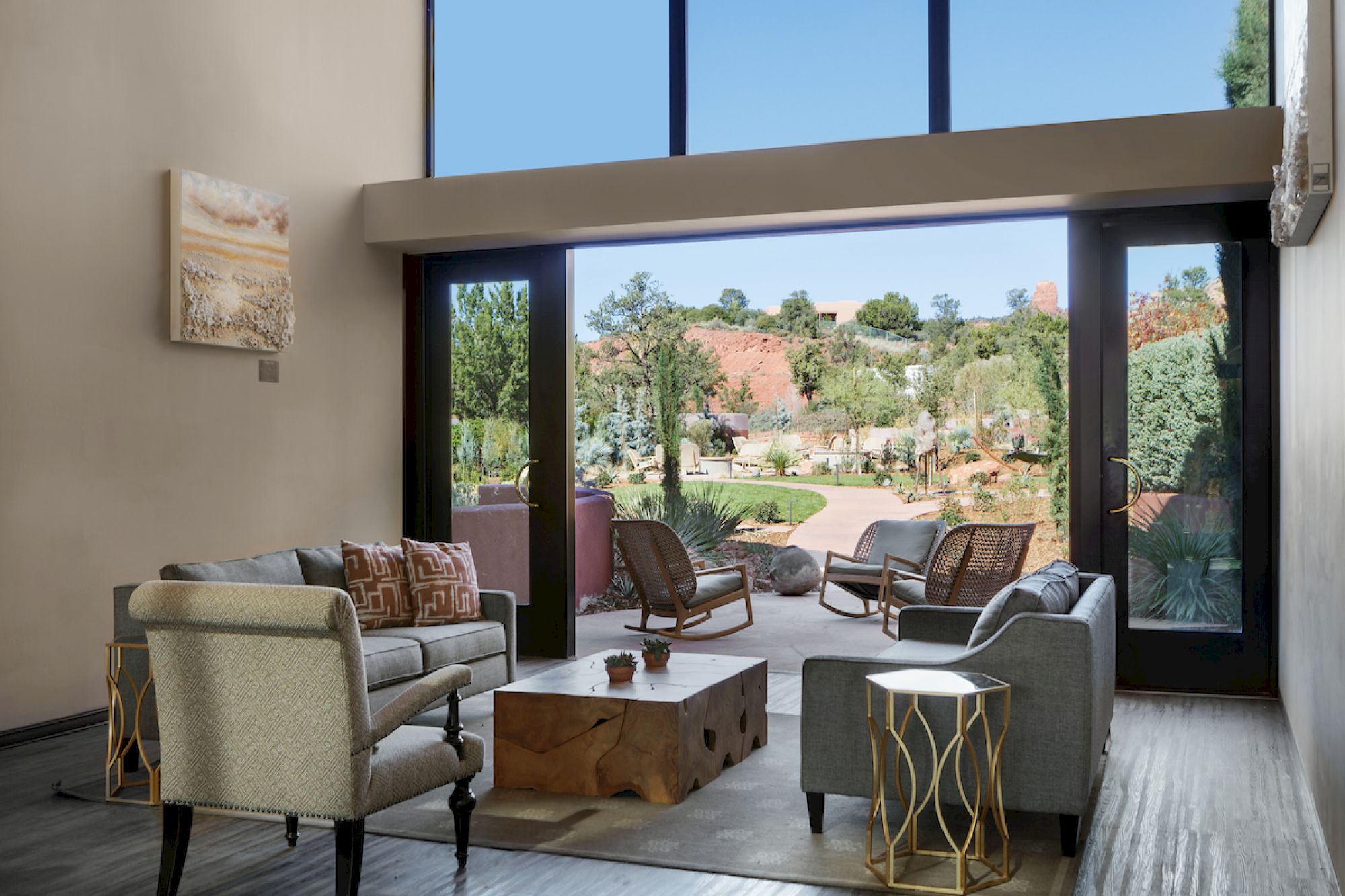 A modern living room with a wooden coffee table, chairs, sofas, artworks, and large windows opening to a garden with outdoor seating and a lush view.