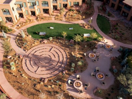 The image shows an aerial view of a courtyard with a labyrinth, seating areas with fire pits, greenery, pathways, and adjacent buildings.