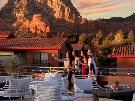 Four people are taking a selfie on a rooftop patio with a beautiful mountainous landscape in the background at sunset.