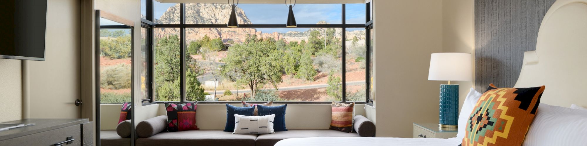 A modern bedroom featuring a large bed, colorful pillows, a cozy window seat, dresser, TV, and expansive windows offering a scenic outdoor view.