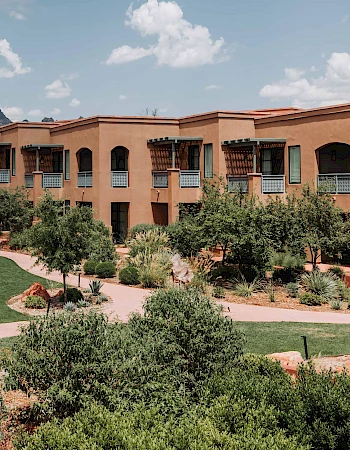 The image showcases a resort or hotel building with terracotta-colored walls, surrounded by green landscaping, paths, and a mountainous backdrop.