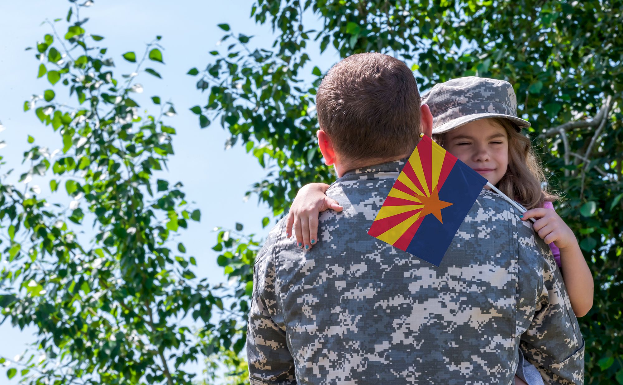 A soldier in camouflage uniform hugs a child wearing a matching hat, with an Arizona flag visible, trees in the background.