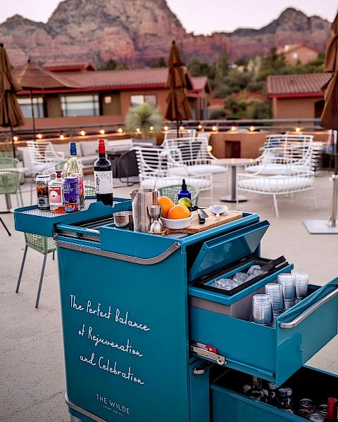 A bar cart with drinks and ingredients sits on a patio with seating, against a backdrop of red rock mountains in the distance.