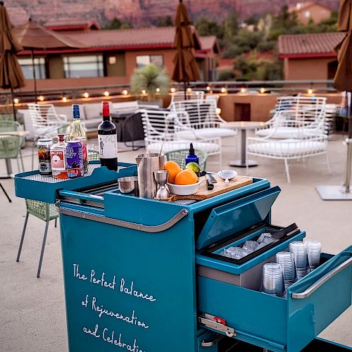 A bar cart with drinks and ingredients sits on a patio with seating, against a backdrop of red rock mountains in the distance.