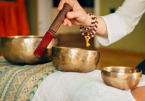 A person is using a mallet to play Tibetan singing bowls placed on a cloth-covered surface, likely for a sound healing session.