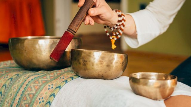 A person is using a mallet to play Tibetan singing bowls placed on a cloth-covered surface, likely for a sound healing session.