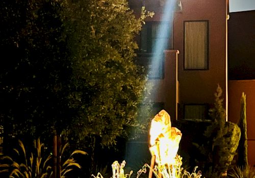 The image shows a nighttime scene with a bright light illuminating plants and sculptures in front of a building under a full moon.