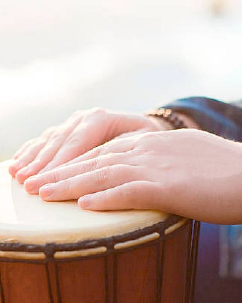 Hands adorned with beaded bracelets are playing a djembe drum in an outdoor setting.